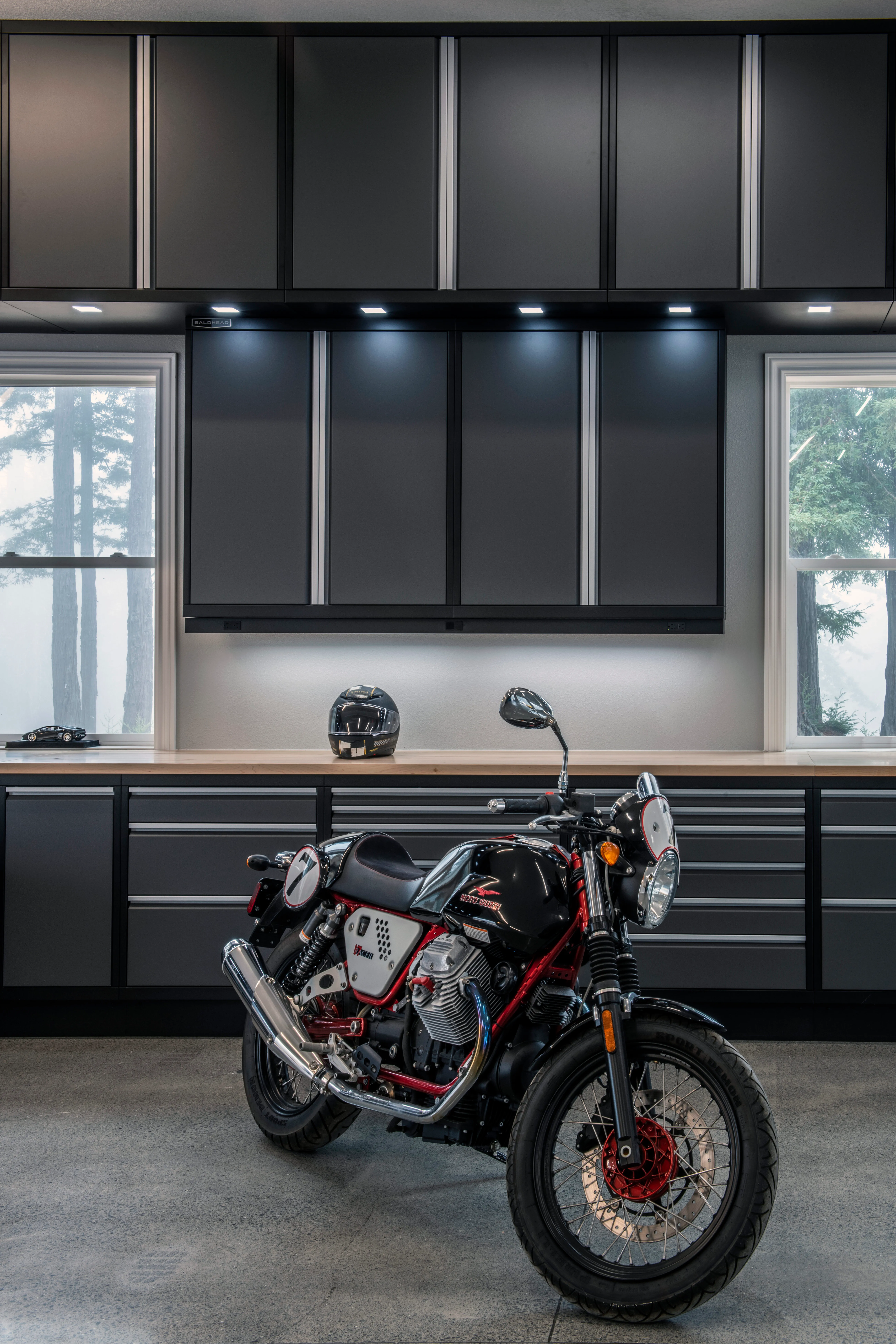 Premium garage cabinet setup by Baldhead Cabinets, featuring matte black and gray powder-coated steel cabinetry with LED task lighting, butcher block countertop, and a classic Moto Guzzi V7 motorcycle parked in a high-end garage workspace.