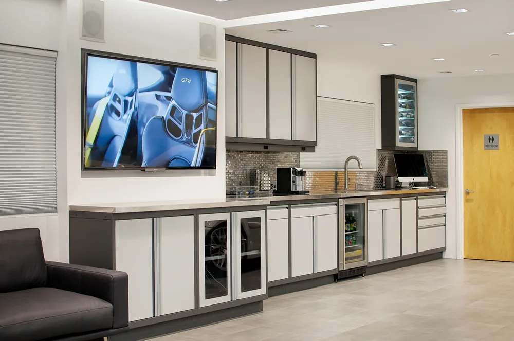 Modern lounge kitchenette featuring Baldhead Cabinets in a two-tone powder-coated finish, integrated beverage cooler, under-cabinet lighting, tile backsplash, wall-mounted TV, and a sleek coffee station, ideal for luxury office or showroom environments.