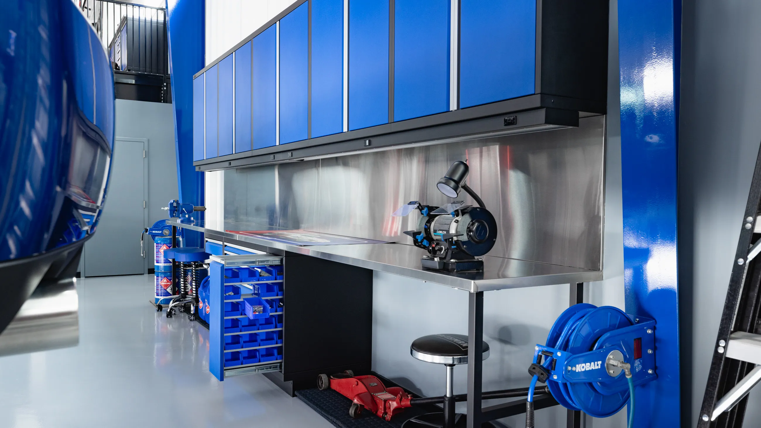 Premium garage workstation featuring custom Baldhead Cabinets in blue with aluminum trim. Stainless steel countertop and backsplash provide a durable, professional-grade work surface. Integrated tool storage, overhead cabinets, and a mounted hose reel offer efficient garage organization. Ideal for automotive shops and home garages seeking high-end cabinetry solutions.