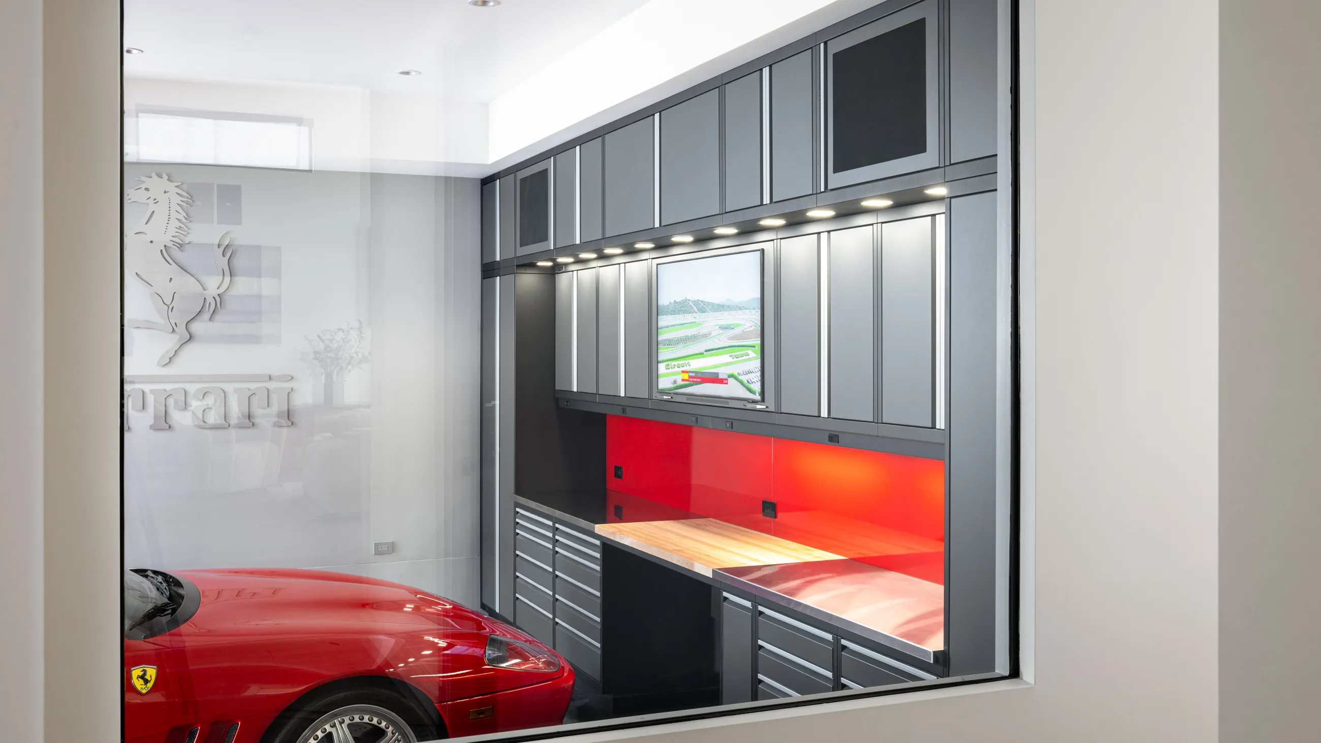 Luxury garage cabinetry setup by Baldhead Cabinets featuring sleek gray and black modular storage, vibrant red backsplash, integrated LED lighting, and a Ferrari parked beside the workspace—ideal for car enthusiasts seeking custom garage organization with high-end aesthetics.