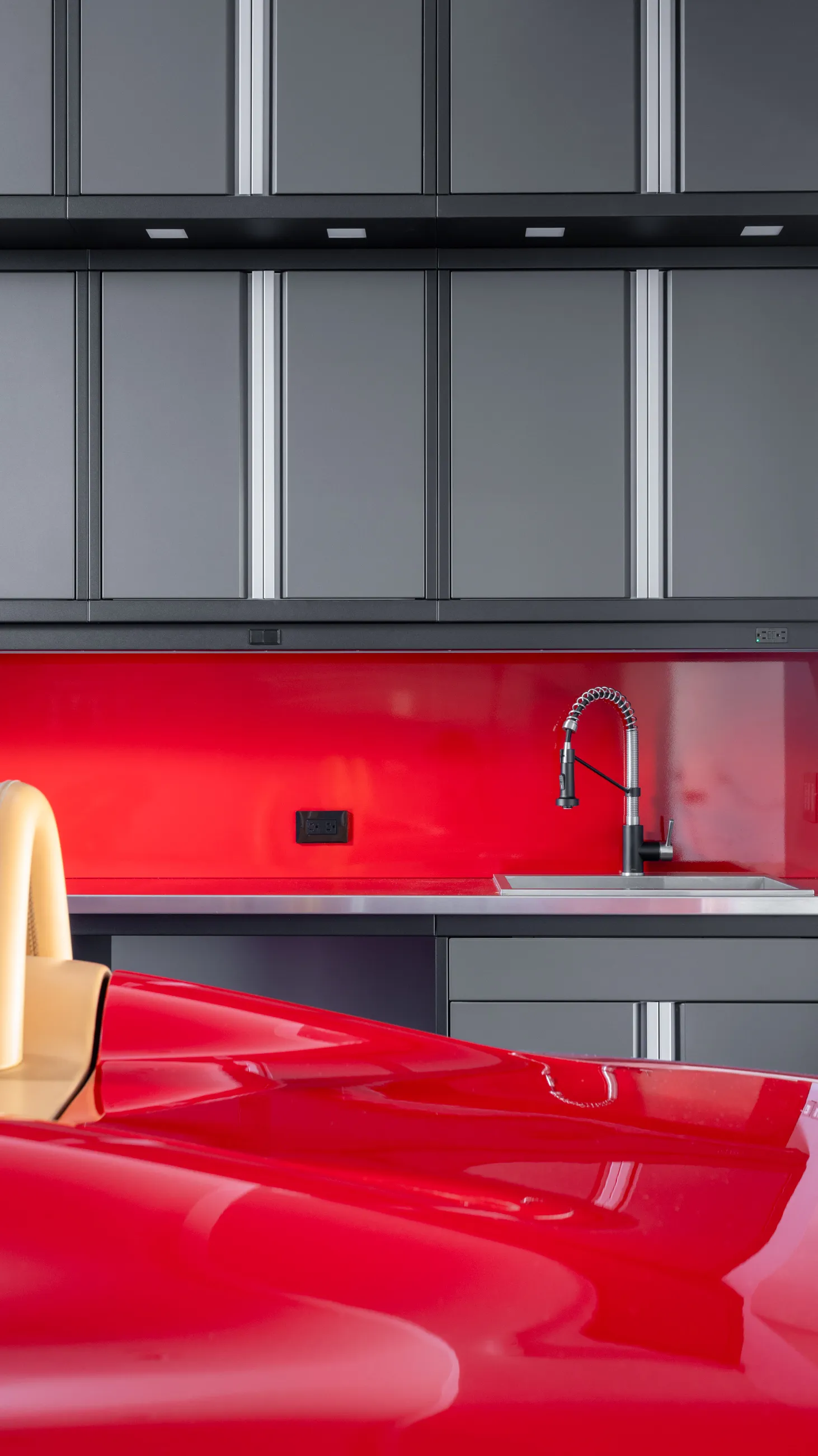 Custom garage setup featuring matte gray Baldhead Cabinets, vivid red backsplash, industrial-style faucet with stainless steel sink, and the rear view of a luxury red sports car, showcasing high-end garage functionality and design.