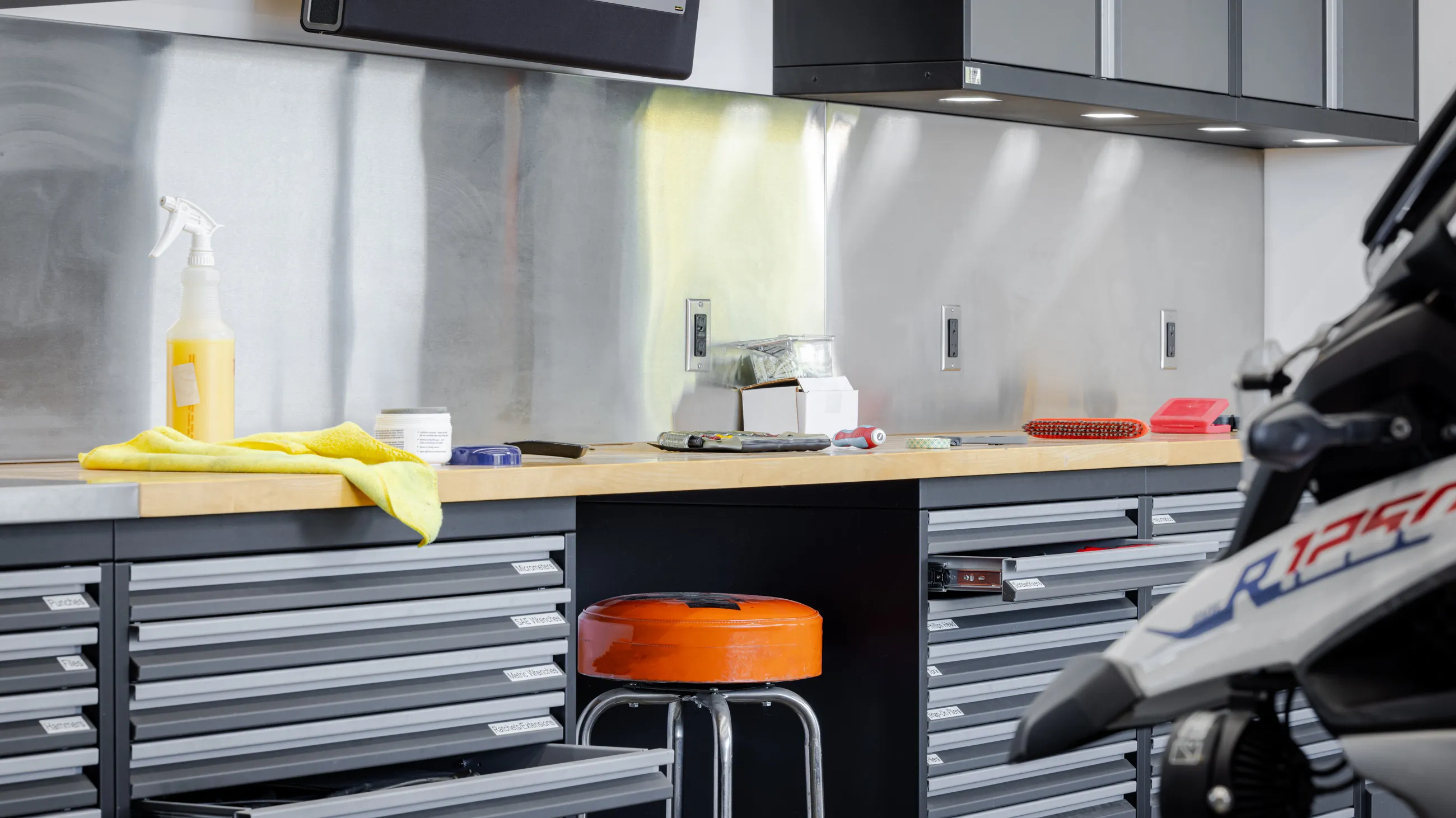 High-performance garage workstation showcasing Baldhead Cabinets’ heavy-duty tool drawers with custom labels, integrated under-cabinet lighting, and solid wood countertops. Stainless steel backsplash and a motorcycle in the foreground emphasize a premium, organized environment for mechanics and riders demanding superior garage storage solutions.