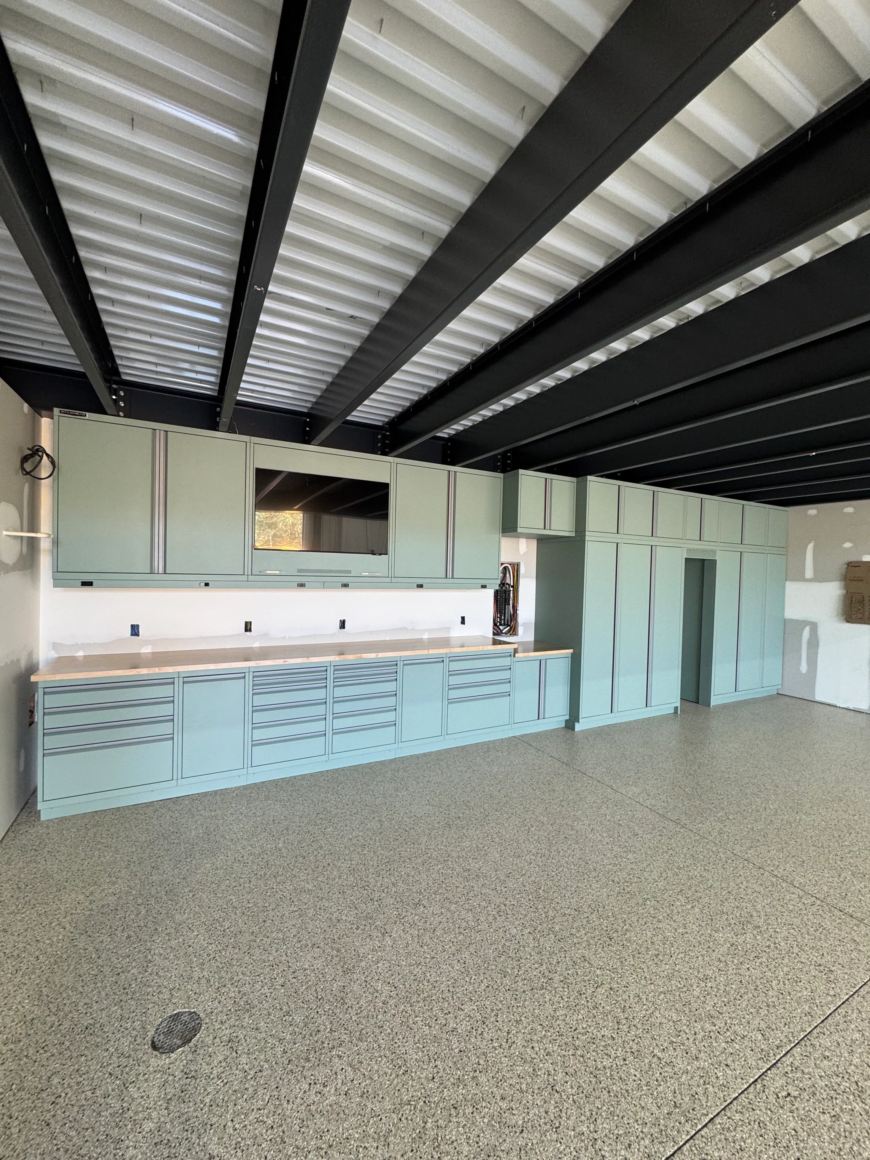 Modern custom garage cabinets by Baldhead Cabinets in soft green powder-coated finish, featuring integrated workspace, full-height storage, ample drawers, butcher block countertops, and epoxy-coated flooring beneath exposed steel beam ceiling.