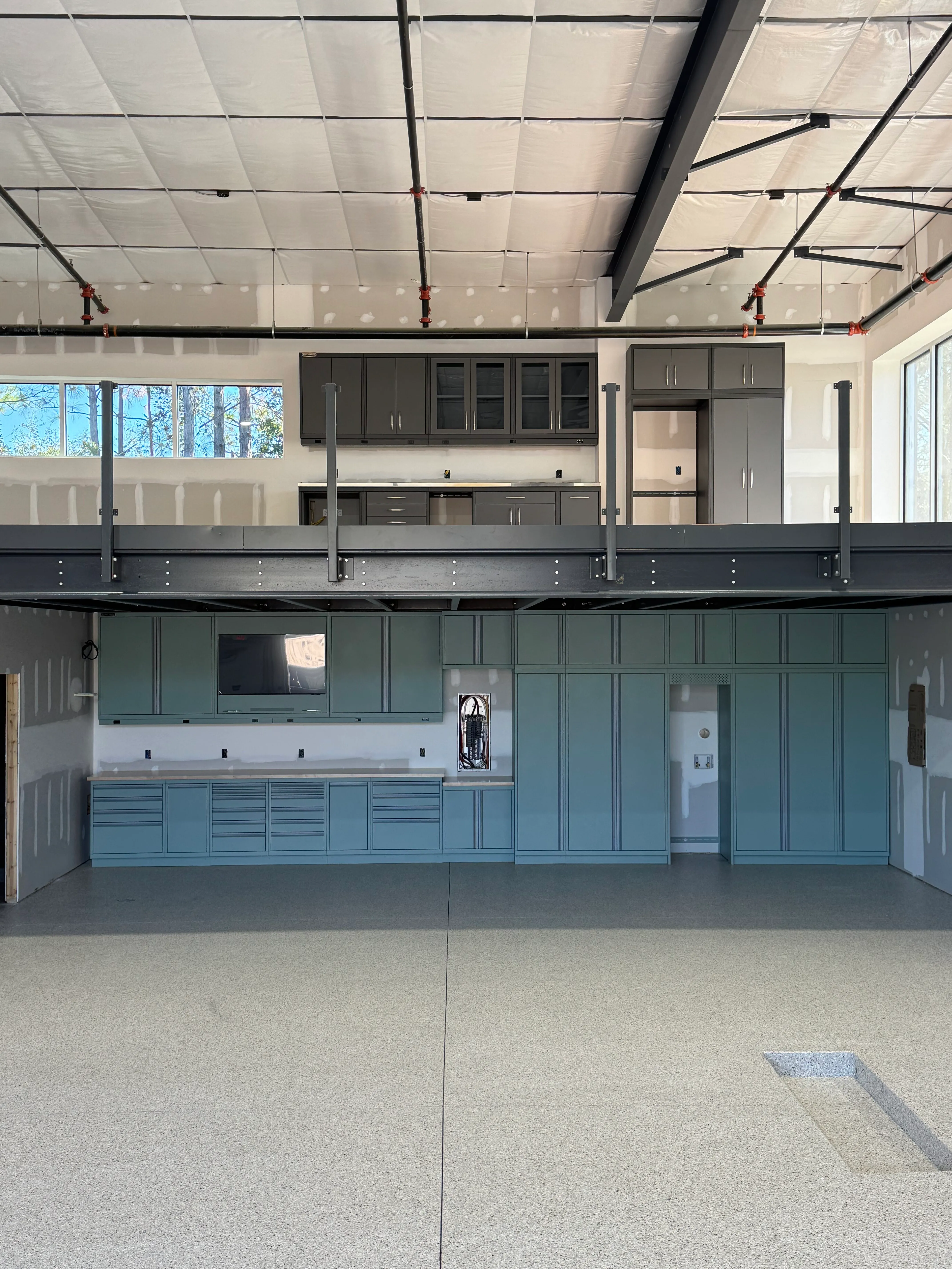 Dual-level custom garage by Baldhead Cabinets featuring soft green lower cabinetry with integrated workspace and TV mount, paired with upper mezzanine dark gray cabinets, epoxy flooring, and exposed steel structure in a modern industrial workshop design.