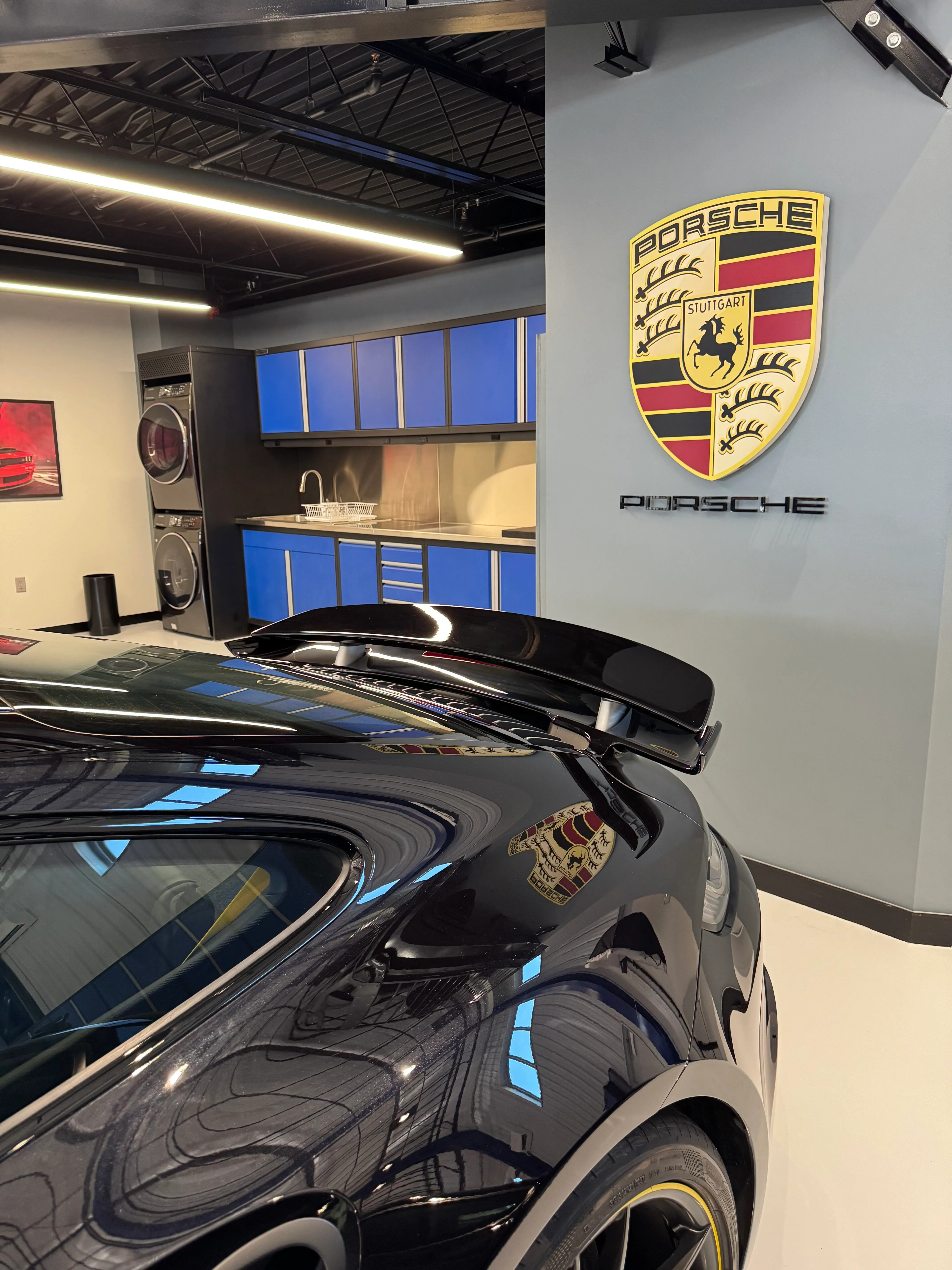 Luxury garage featuring custom blue Baldhead Cabinets and stainless steel countertops, perfectly complementing a high-performance Porsche. Premium garage design with built-in sink, laundry setup, and sleek storage solution for automotive enthusiasts.