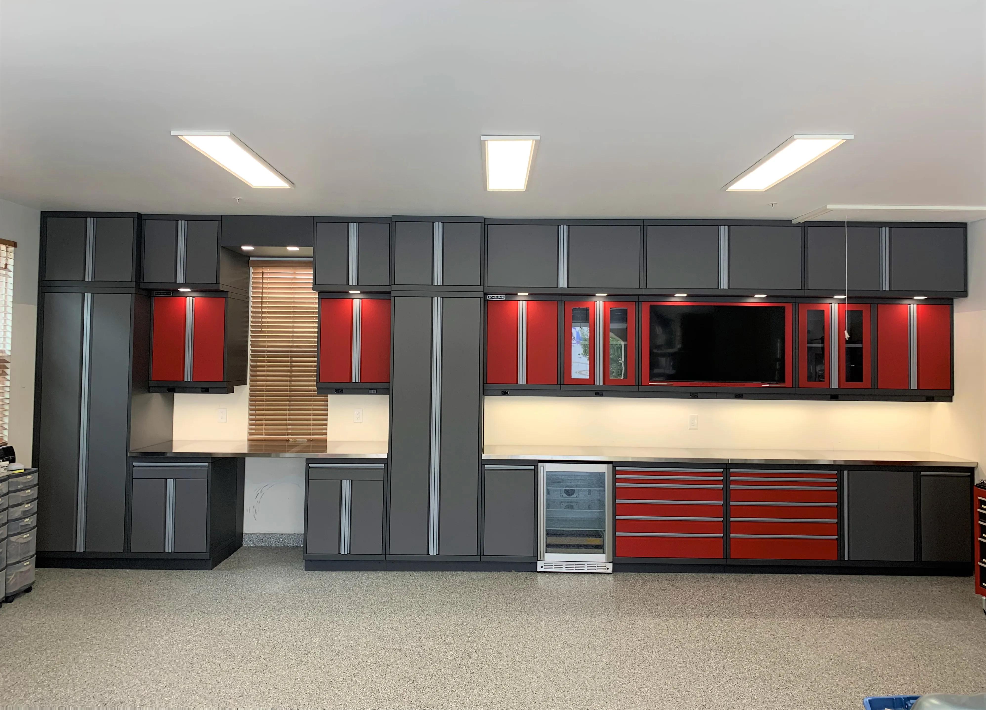 Custom garage cabinetry by Baldhead Cabinets in a bold red and charcoal finish, featuring upper and lower modular storage, integrated beverage cooler, under-cabinet lighting, workstations, and a wall-mounted TV, all set against a speckled epoxy floor.