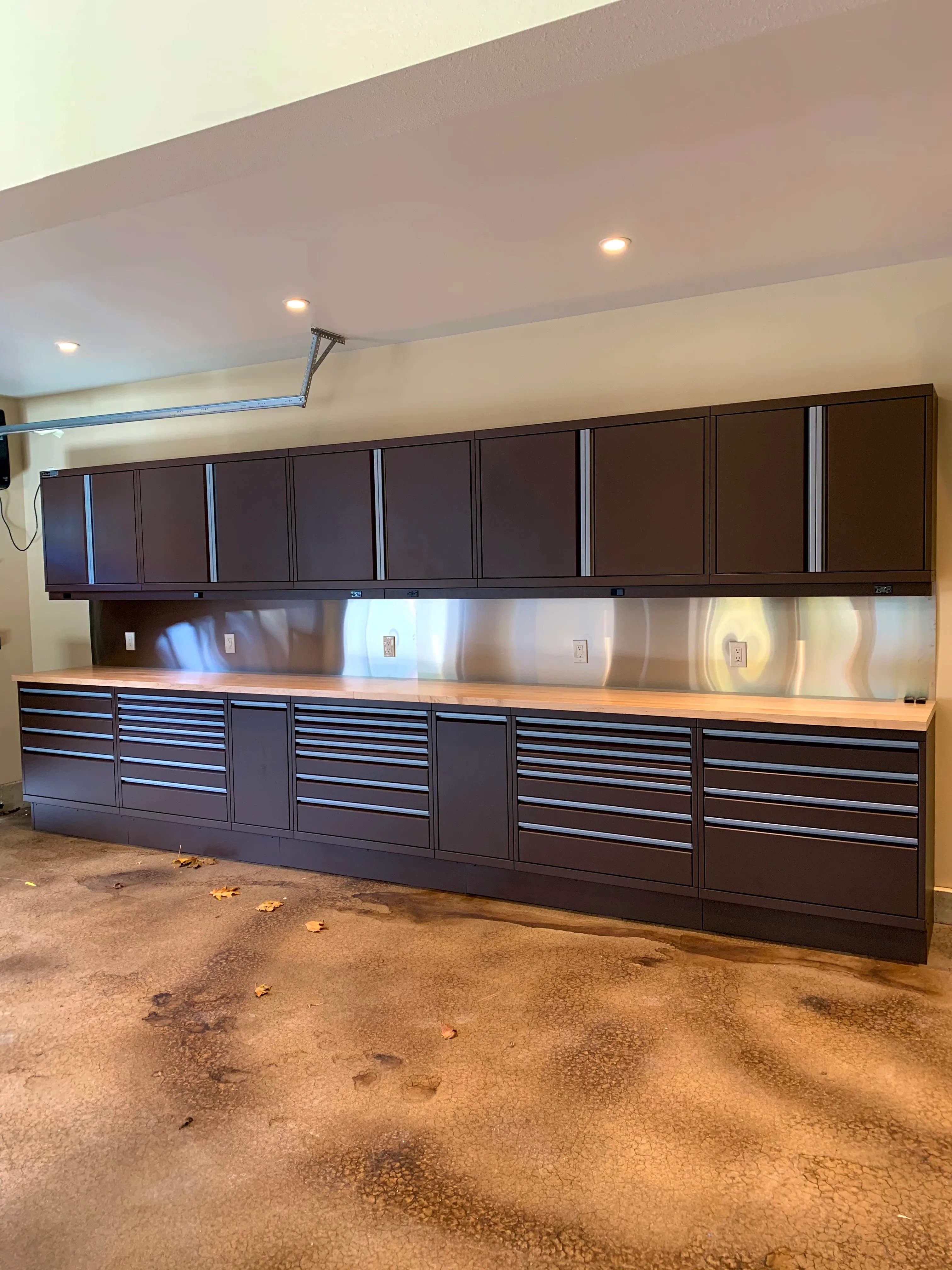 Custom brown powder-coated garage cabinets by Baldhead Cabinets featuring full-length upper storage, stainless steel backsplash, butcher block countertop, and wide tool drawers, set against a rustic stained concrete floor in a premium residential garage.
