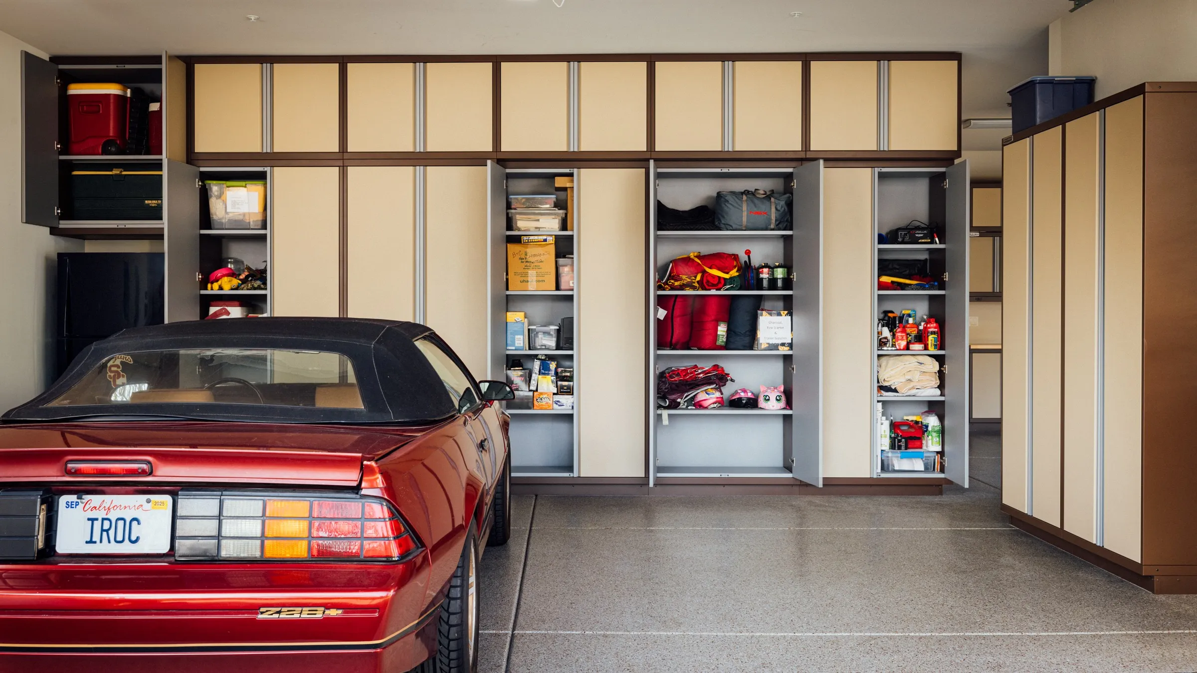 Spacious garage with custom Baldhead Cabinets in tan and brown finish, showcasing open storage filled with tools, gear, and household items, alongside a vintage red Camaro parked on a polished epoxy floor.