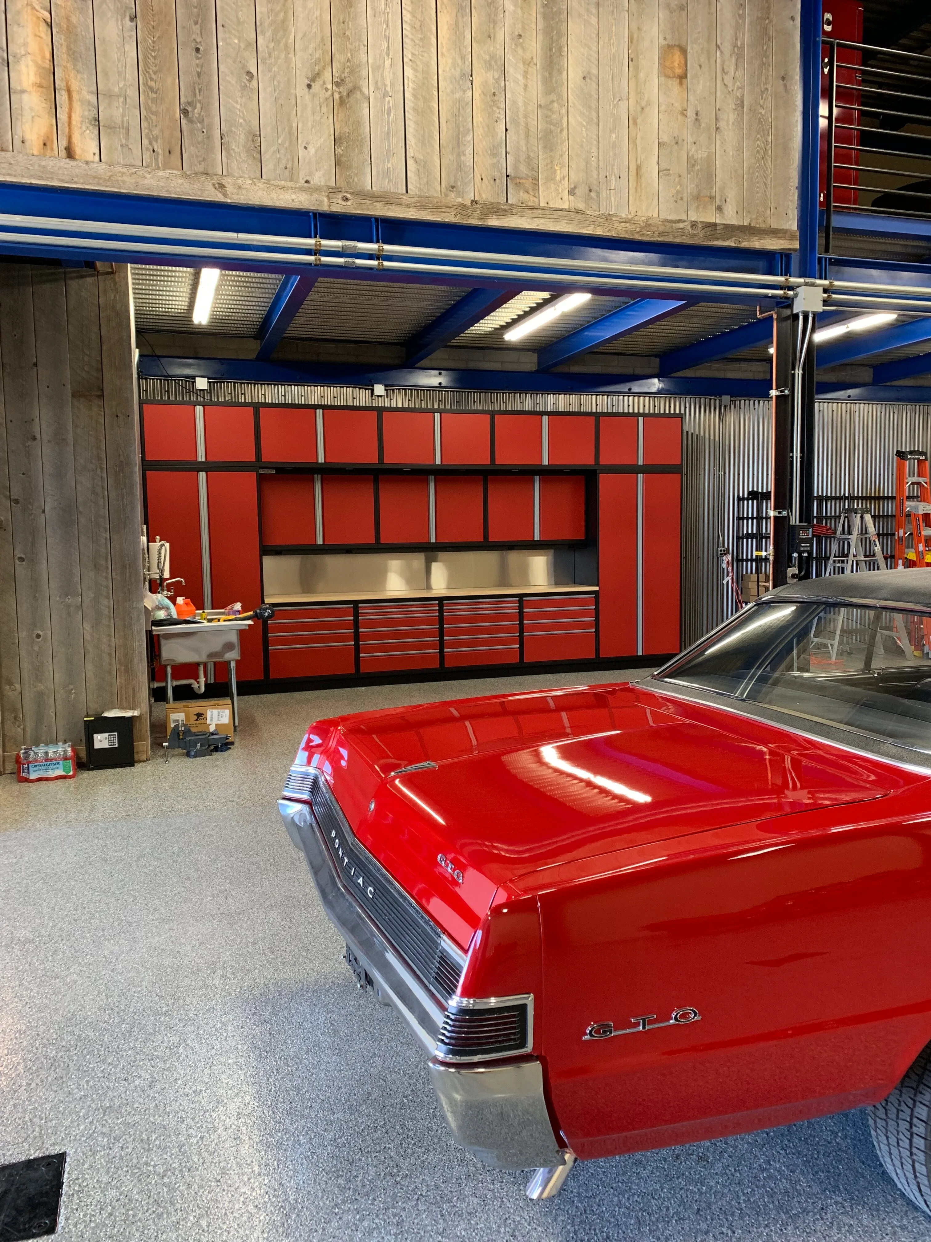 Custom red and black garage cabinetry by Baldhead Cabinets with stainless steel backsplash, paired with a classic red Pontiac GTO and rustic wood and corrugated metal finishes, set in a professional-grade automotive shop with epoxy flooring.