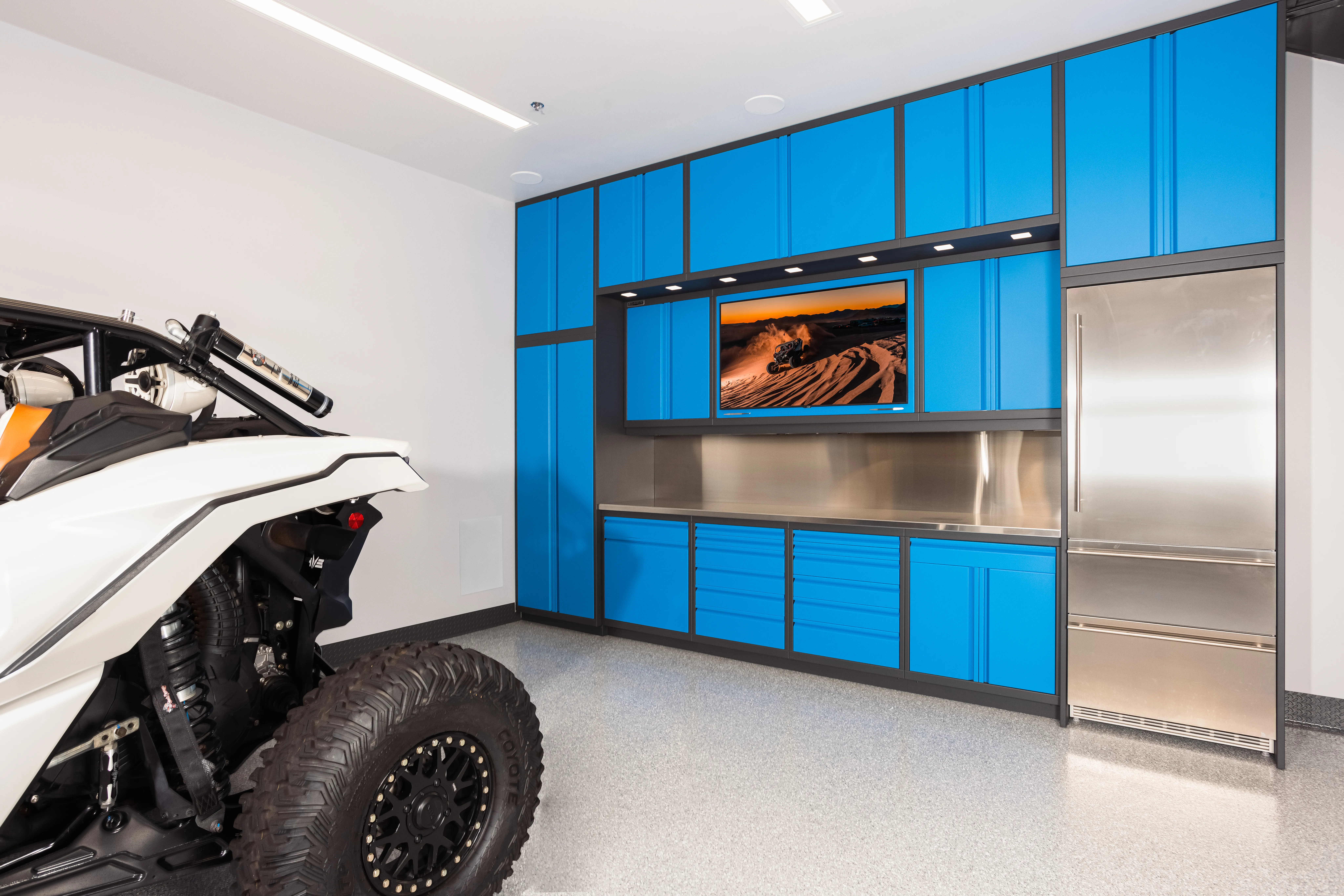 Bold blue custom garage cabinets by Baldhead Cabinets with integrated TV, stainless steel countertops and refrigerator, under-cabinet lighting, and heavy-duty drawers, paired with a white off-road vehicle in a high-performance motorsports workspace.