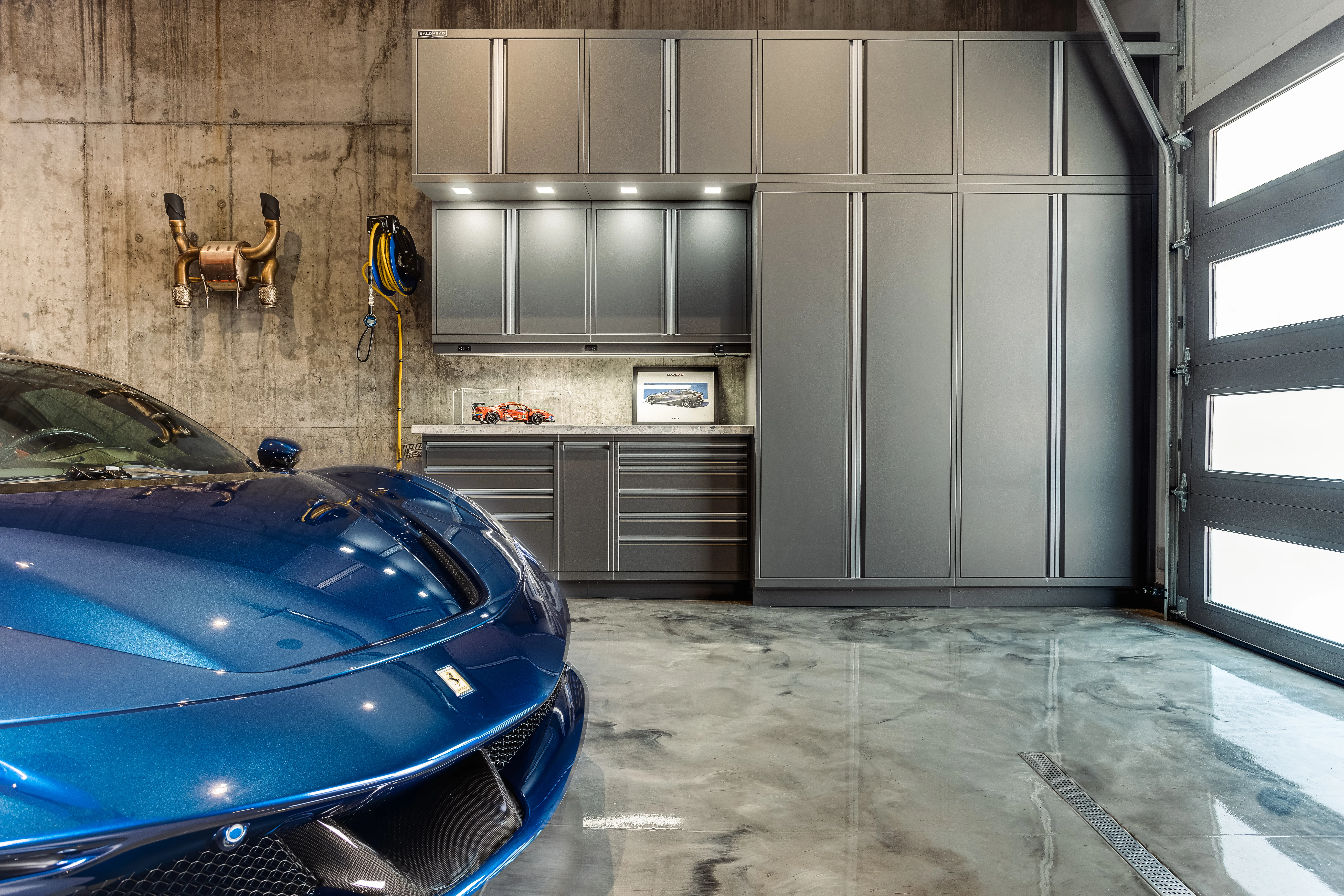 Luxury garage setup featuring matte metallic Baldhead Cabinets with integrated LED lighting, concrete accent wall, polished epoxy flooring, and a deep blue Ferrari supercar, showcasing premium automotive storage and design excellence.
