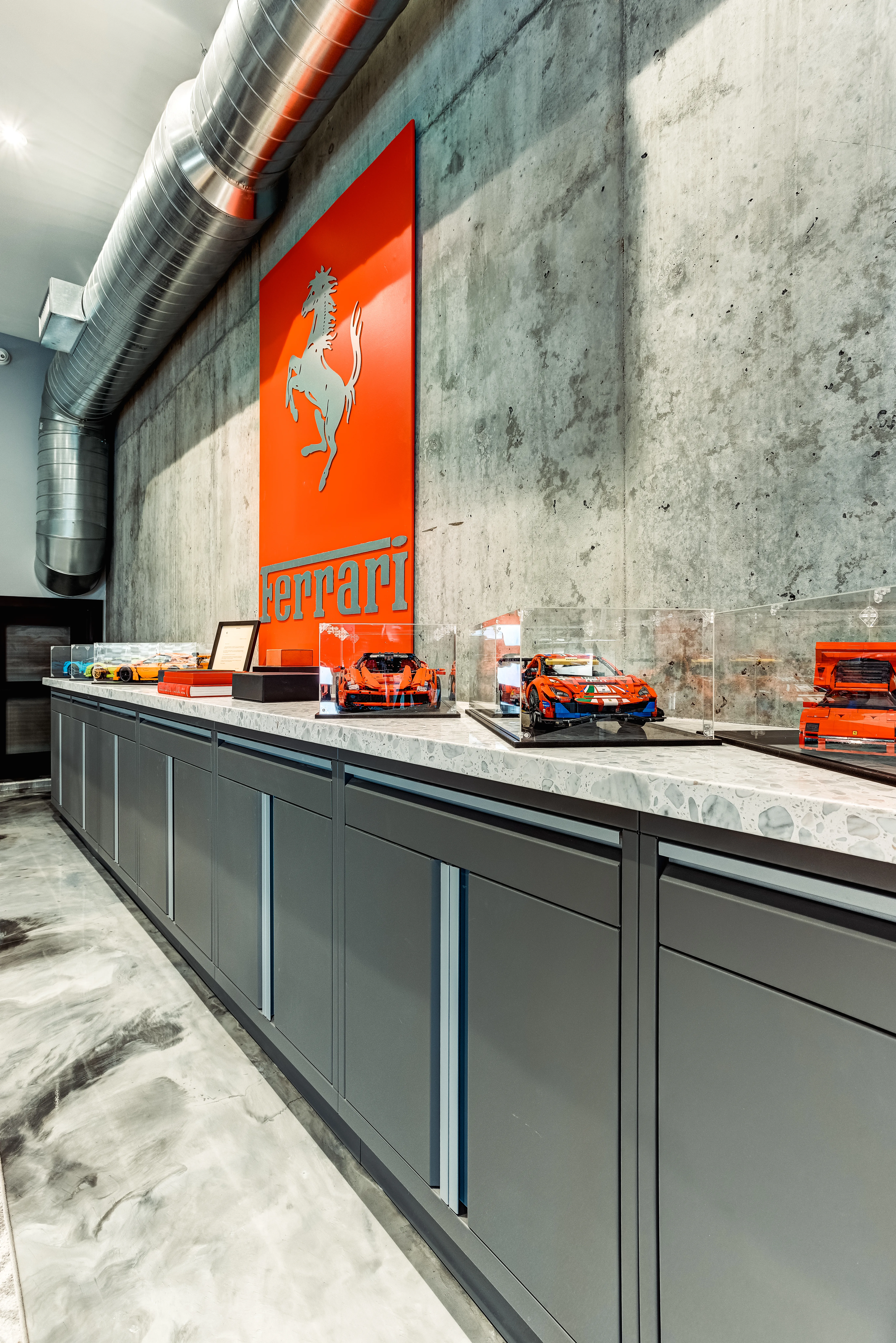 Custom Baldhead Cabinets installation in a luxury garage featuring sleek matte gray cabinetry with integrated handles, a durable stone-patterned countertop, and a Ferrari-themed display wall — perfect for automotive enthusiasts seeking high-end garage organization solutions.
