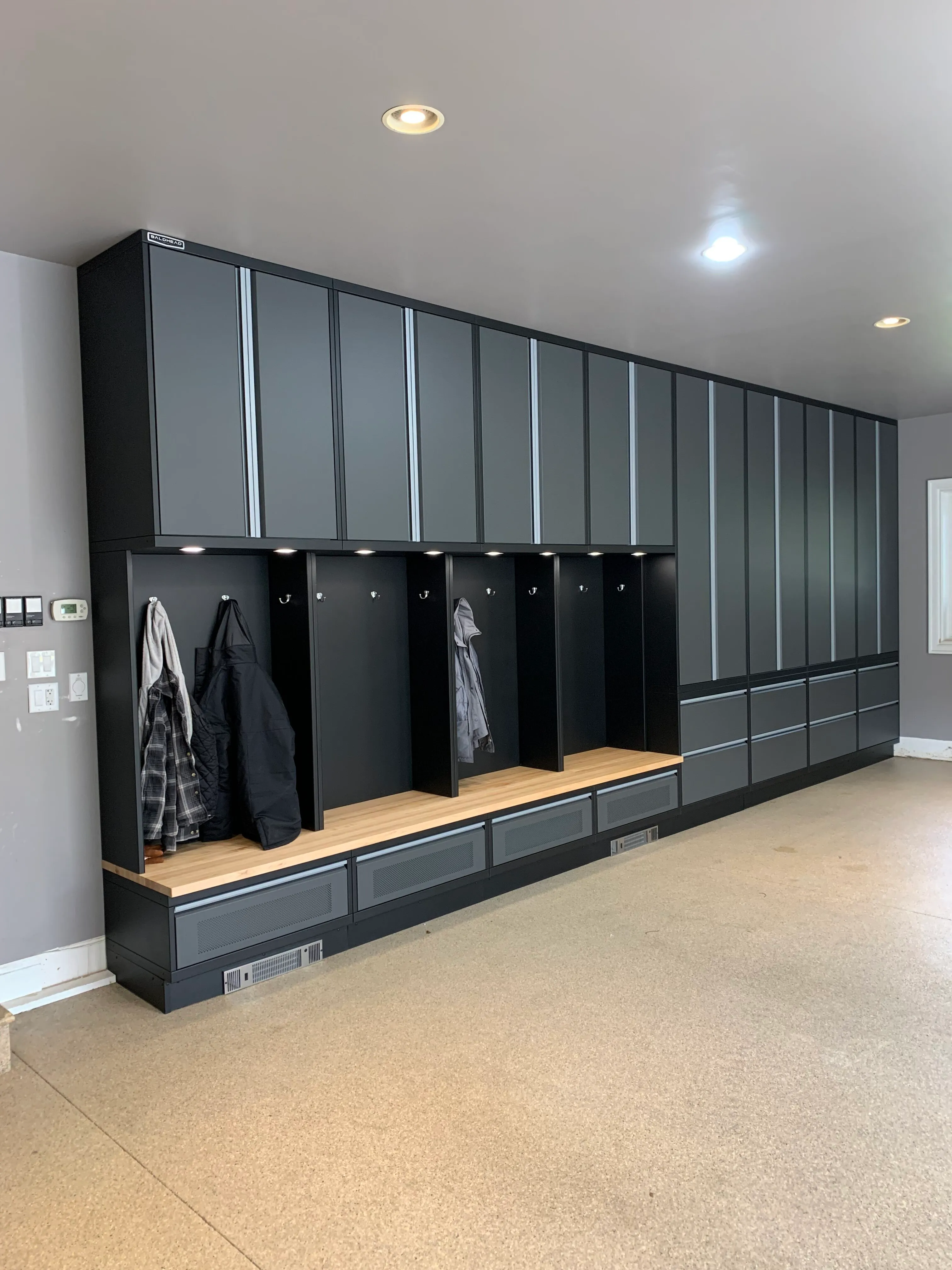 Custom mudroom locker system by Baldhead Cabinets featuring tall matte black and gray cabinets, individual cubby storage with coat hooks, integrated bench seating with wood tops, and under-bench ventilation in a high-end residential entryway.