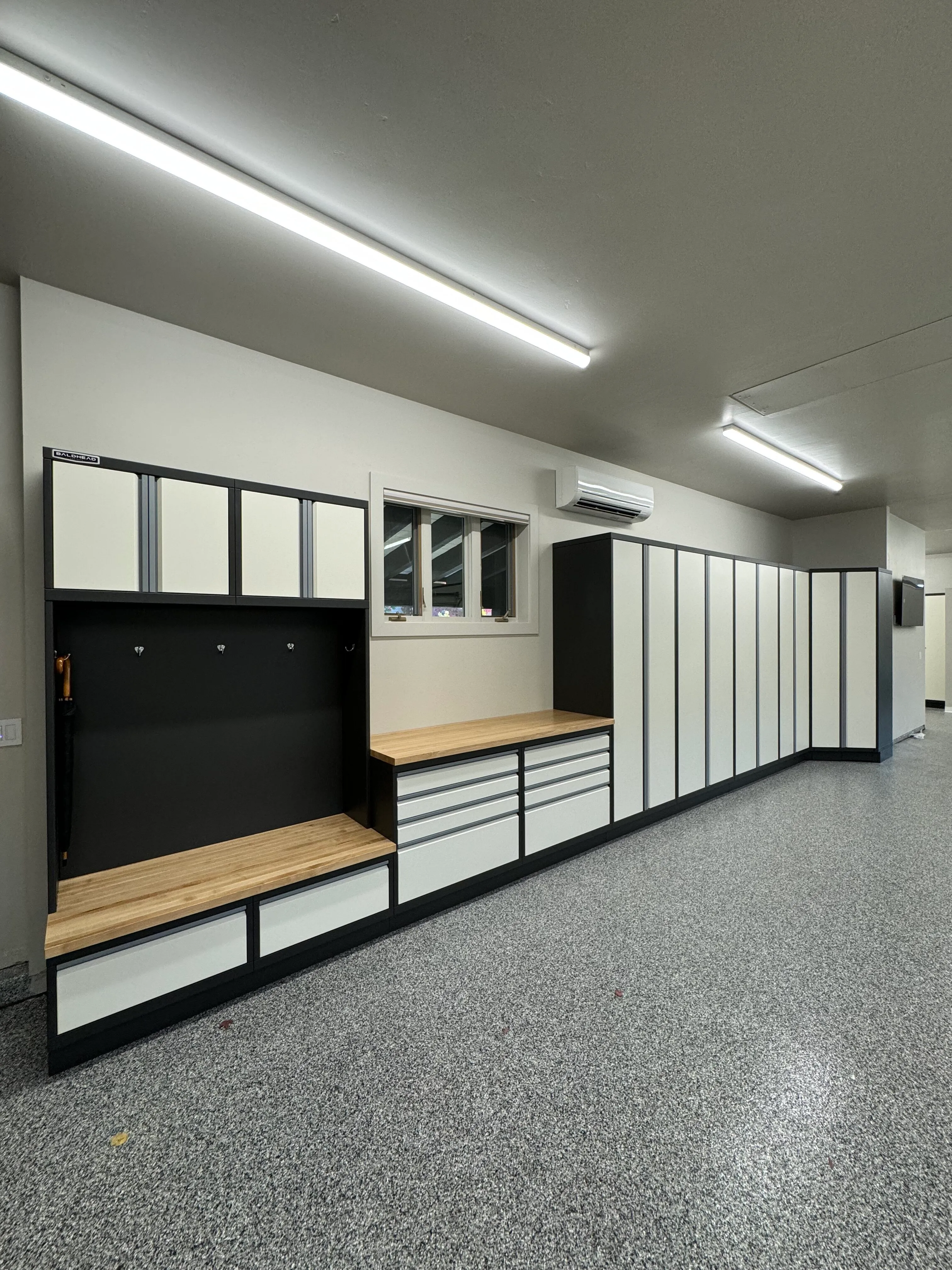 Custom garage entry system by Baldhead Cabinets featuring two-tone powder-coated cabinetry in cream and black, integrated locker bench with butcher block seating, ample drawer storage, full-height cabinets, and speckled epoxy flooring in a climate-controlled space.