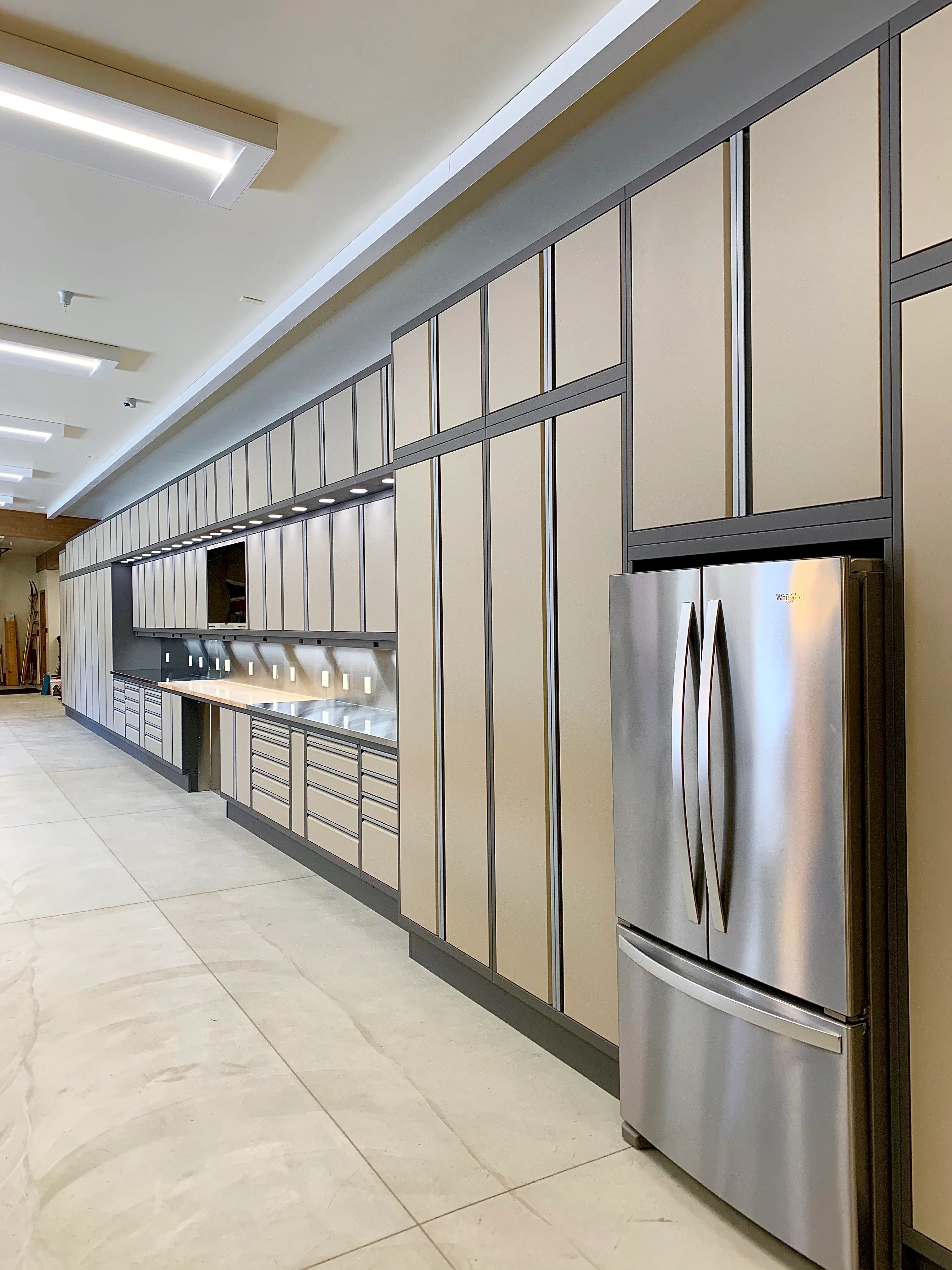 Expansive custom cabinetry by Baldhead Cabinets in a tan and charcoal powder-coated finish, featuring full-length vertical storage, integrated lighting, stainless steel work surfaces, and built-in Whirlpool refrigerator in a luxury garage or workshop space.