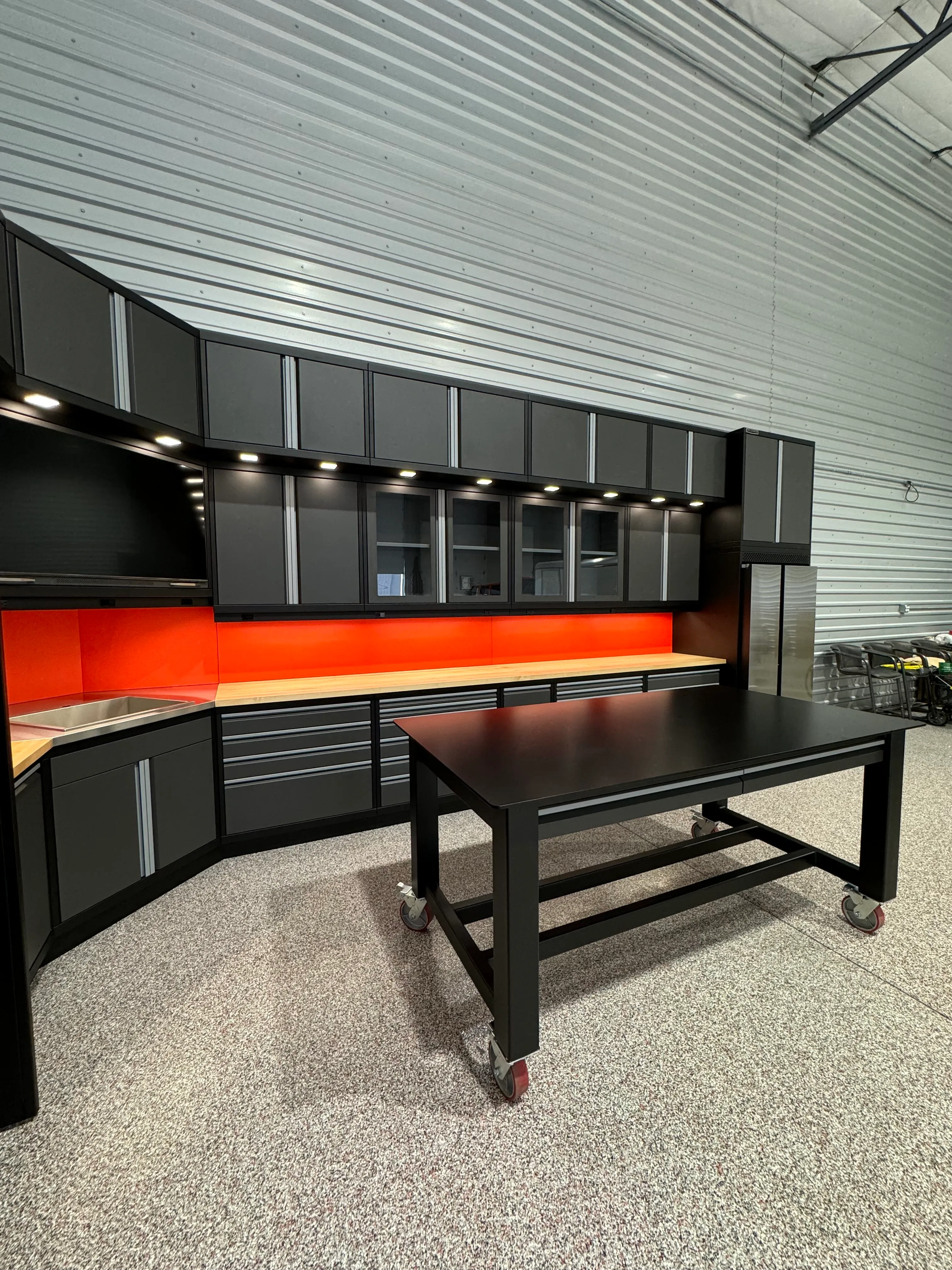 Custom industrial garage setup by Baldhead Cabinets featuring matte black and gray powder-coated cabinetry, bold red backsplash, butcher block countertops, integrated lighting, stainless steel sink, and a heavy-duty rolling workstation on speckled epoxy flooring.