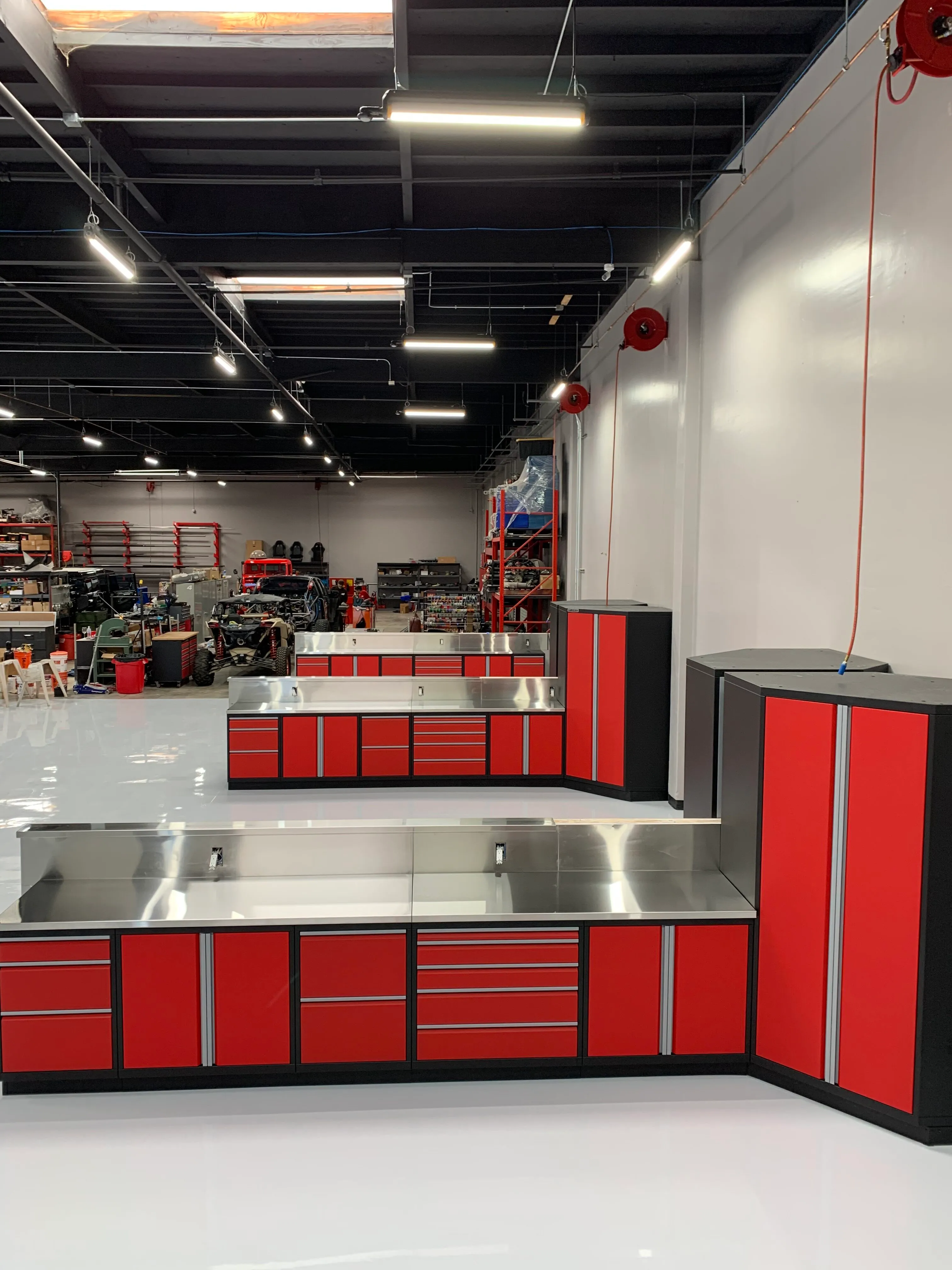 Striking red and black industrial cabinet setup by Baldhead Cabinets featuring modular drawer and door configurations, stainless steel countertops and backsplash, and a high-gloss epoxy floor in a professional-grade automotive workshop.