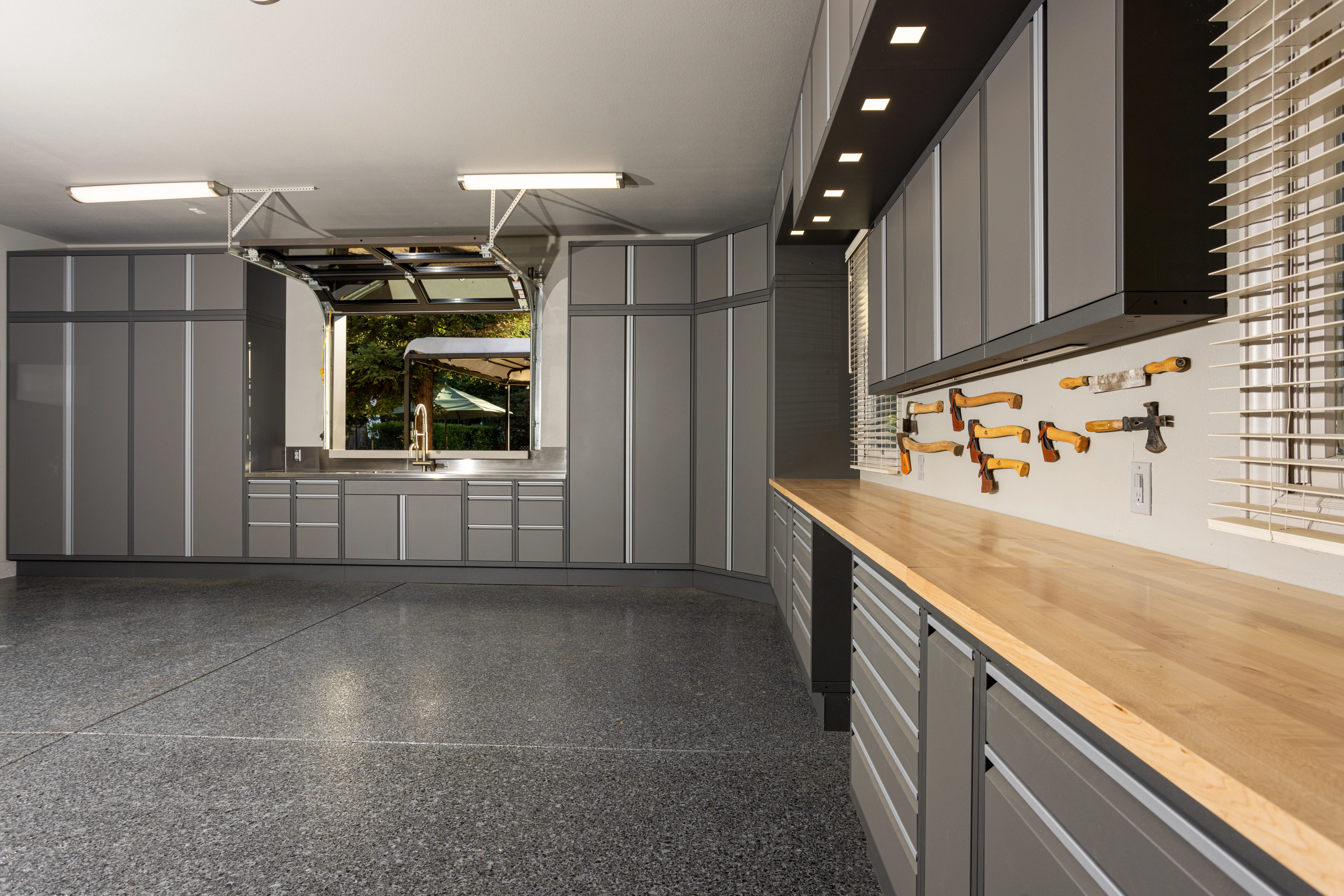 Spacious custom garage by Baldhead Cabinets featuring matte gray powder-coated cabinetry, butcher block countertops, integrated LED task lighting, wall-mounted vintage tools, stainless steel sink, and epoxy-coated flooring for a premium workshop environment.