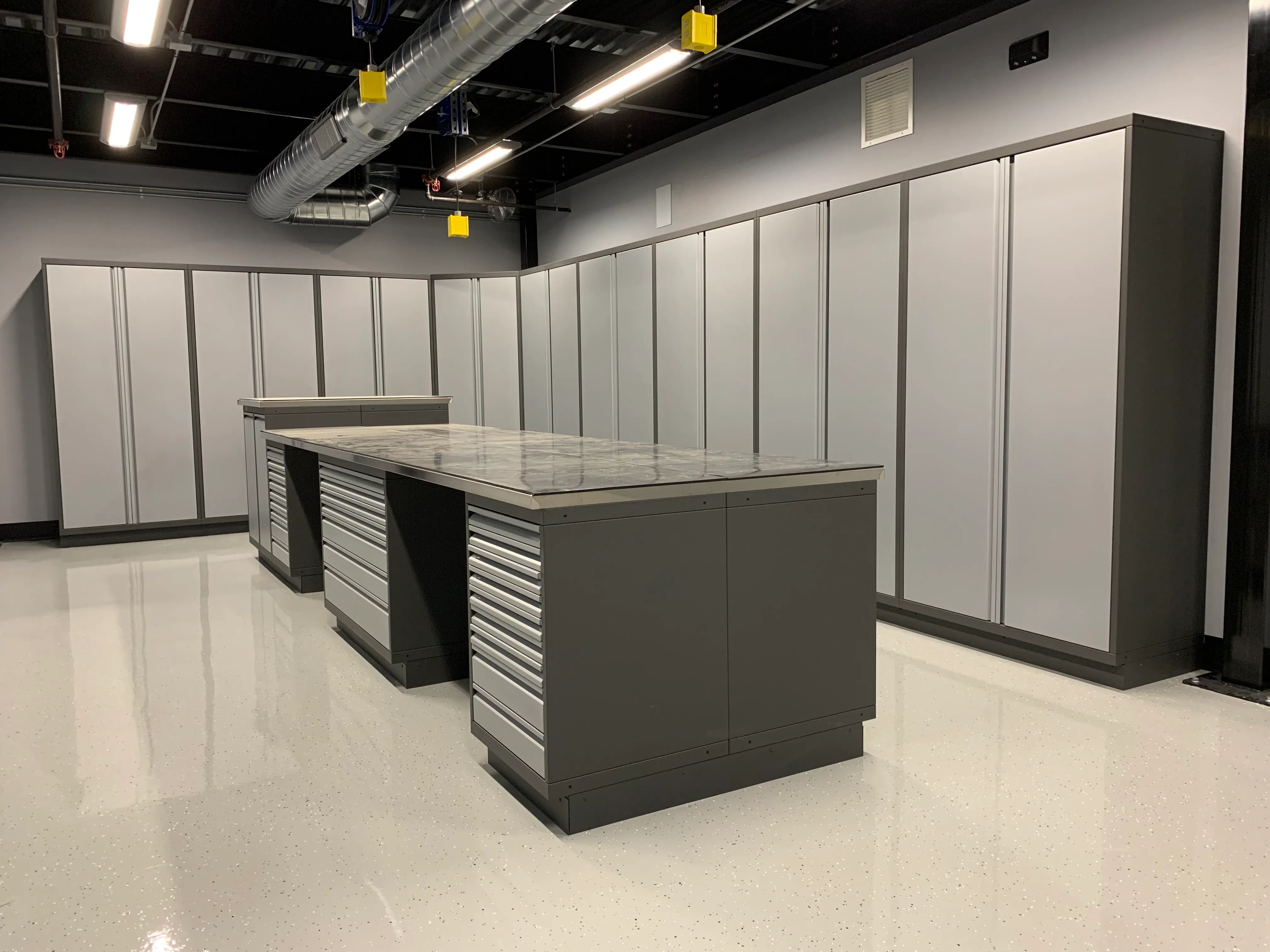 Modern industrial workspace by Baldhead Cabinets featuring matte gray full-height storage cabinets, dual mobile drawer workstations with marble countertops, and a polished speckled epoxy floor beneath exposed ductwork and overhead lighting.