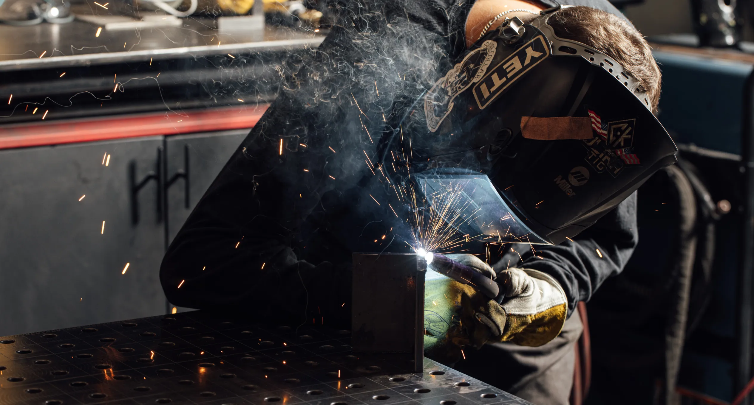 Welder in action, sparks flying while welding a metal piece, wearing welding helmet and gloves.